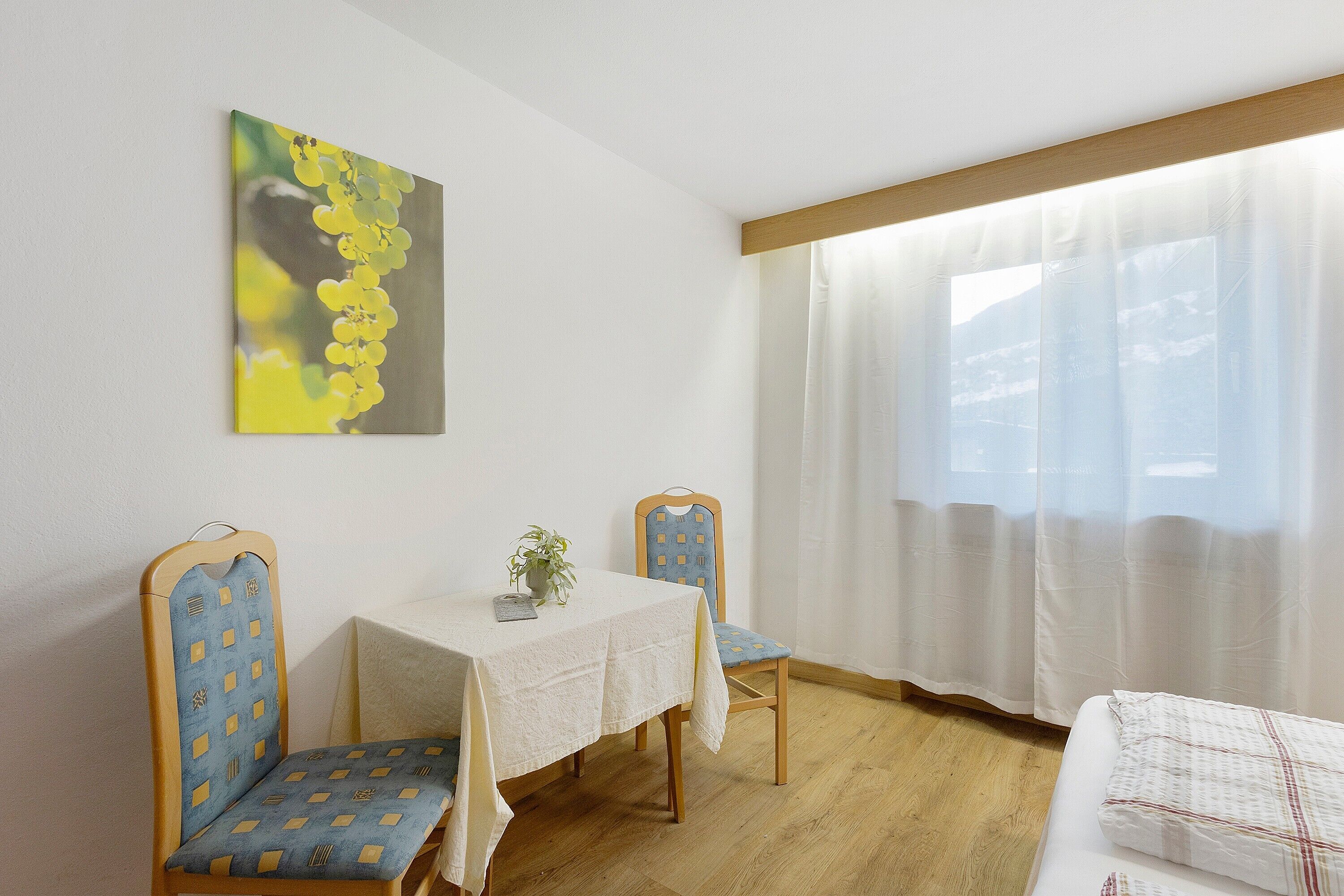 Une pièce avec un lit, une table et une chaise, un tableau au mur et une fenêtre avec des rideaux.