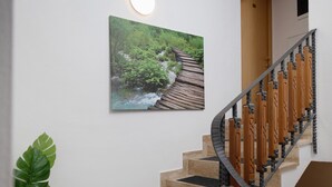 Una escalera con barandas de madera y escalones color beige, un cuadro colgado en la pared que muestra un arroyo boscoso, y una planta en maceta.