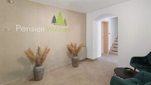 Una zona de recepción con un cartel en la pared que dice 'Pension Waldheim', dos jarrones con plantas secas, una escalera y un sillón verde.
