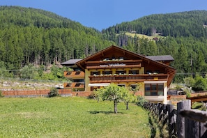 Un edificio de varios pisos con un letrero que dice 'Pension Waldheim', rodeado por una valla de madera, un árbol pequeño y un paisaje montañoso.