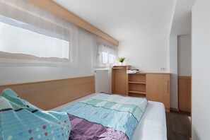 A bedroom with a bed, a window with sheer curtains, a radiator, a small plant, and wooden furniture.