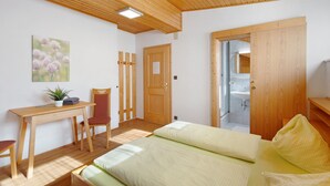 A room with a wooden ceiling, a bed with two pillows, a desk with a chair, a potted plant, and a door leading to a bathroom.