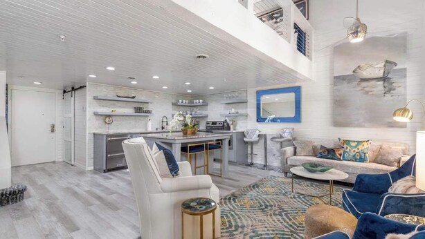 A spacious living area with a high ceiling, a kitchen with white cabinetry, a dining area, and a seating area with blue and white furniture.