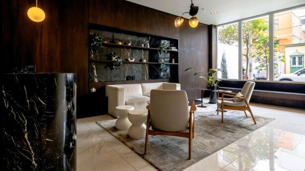A modern lobby with a marble reception desk, a sofa, two armchairs, and a bookshelf with plants.