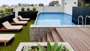 A rooftop pool with lounge chairs, a wooden deck, and city views.
