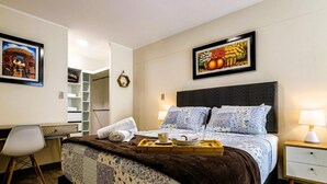 A hotel room with a bed, a desk, a chair, a tray with breakfast items, and two paintings on the walls.