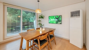 A dining area with a wooden table and chairs, a large window with a view of trees, a wall-mounted TV, and a white air conditioning unit.