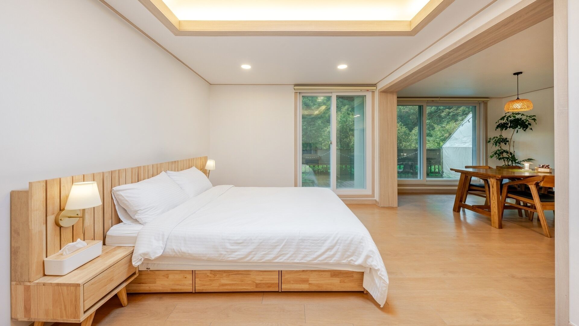 A bedroom with a wooden bed, bedside table, lamp, and a view of the outdoors.