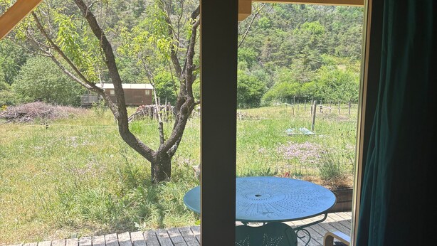 Eine Holzterrasse mit Tisch und Stühlen, Blick auf einen Baum und eine Rasenfläche.