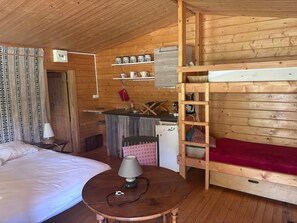 A cozy cabin interior with a bed, a small table with a lamp, a kitchenette with a refrigerator, and a bunk bed.