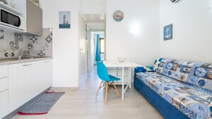 Cuina compacta amb microones, rentatrelle i un rebedor estampat. Es veu una zona de dinar amb una taula blanca i una cadira blava. Un sofà blau amb coixins estampats és en primer pla.