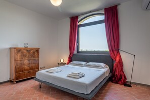 Un dormitorio con cama, cómoda de madera, mesita de noche, lámpara de pie y ventana con cortinas rojas.