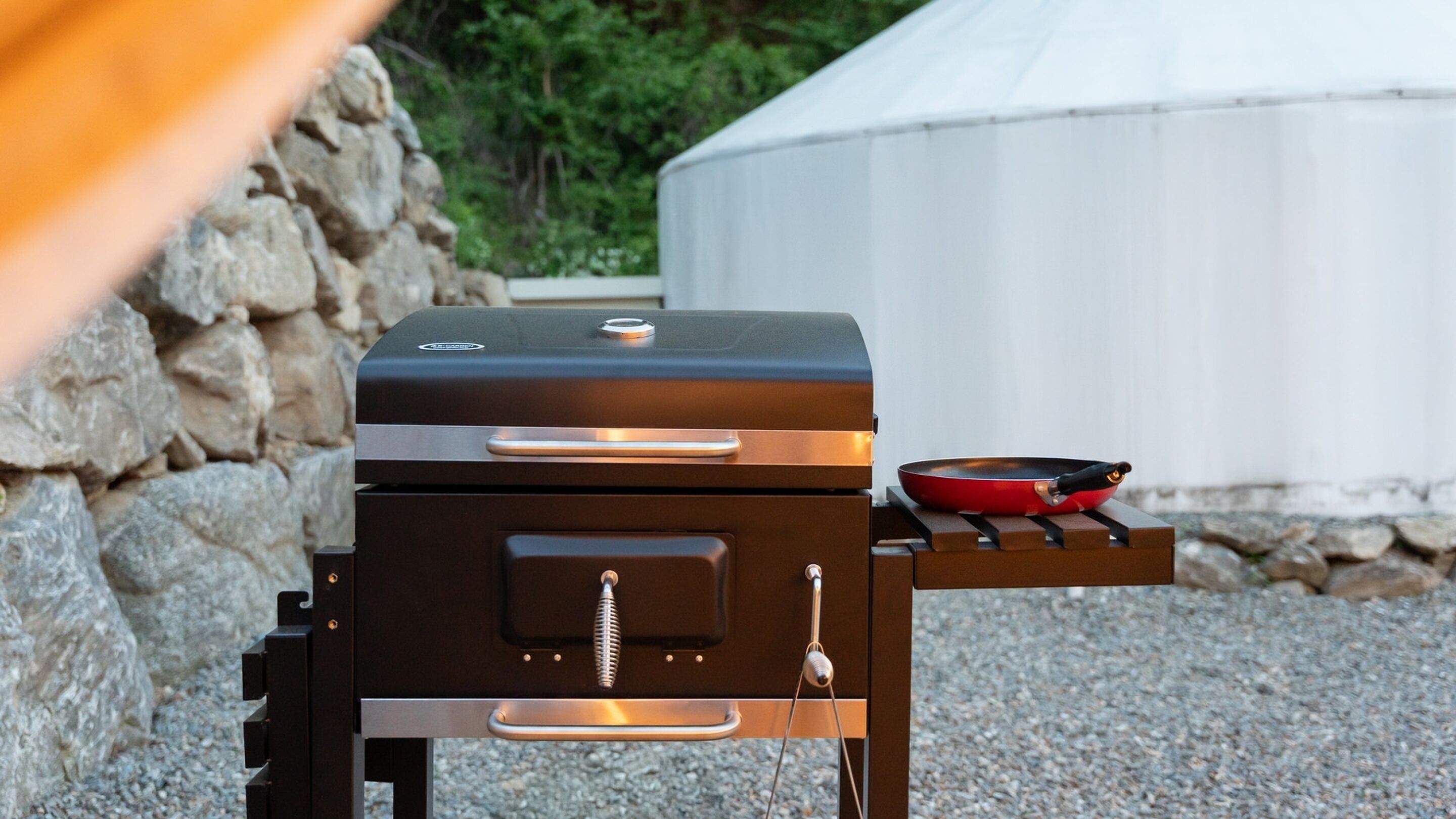 A portable grill with a built-in thermometer and a side burner, set on a gravel surface near a stone wall and a white tent-like structure.