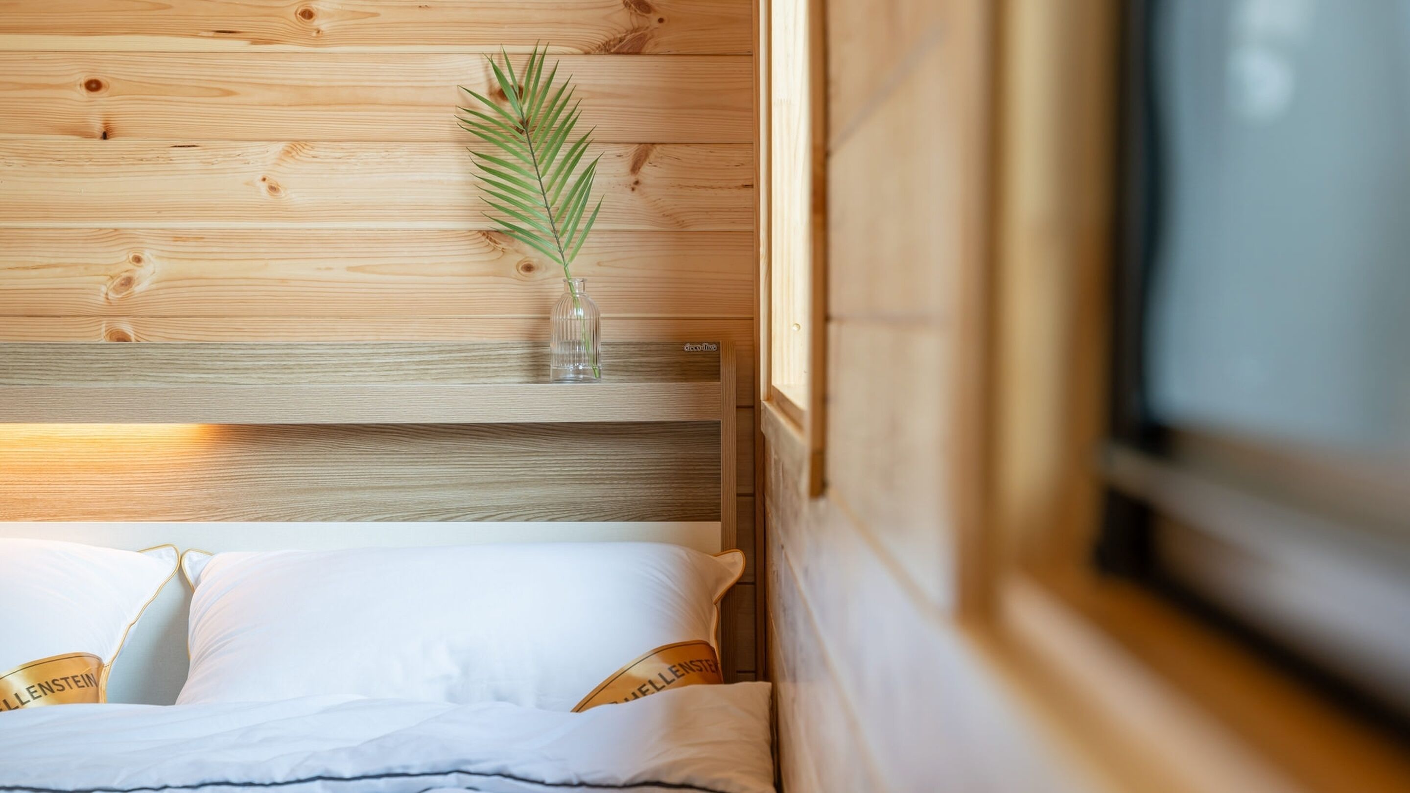 A wooden bed with white bedding and a clear glass vase with a plant on the headboard.