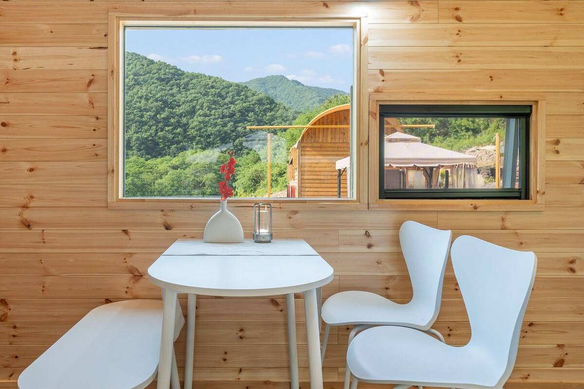 Ein Zimmer in einer Holzhütte mit großem Fenster mit Blick auf einen bewaldeten Berg. Es gibt einen weißen Tisch mit zwei Stühlen und eine Vase mit roten Blumen.