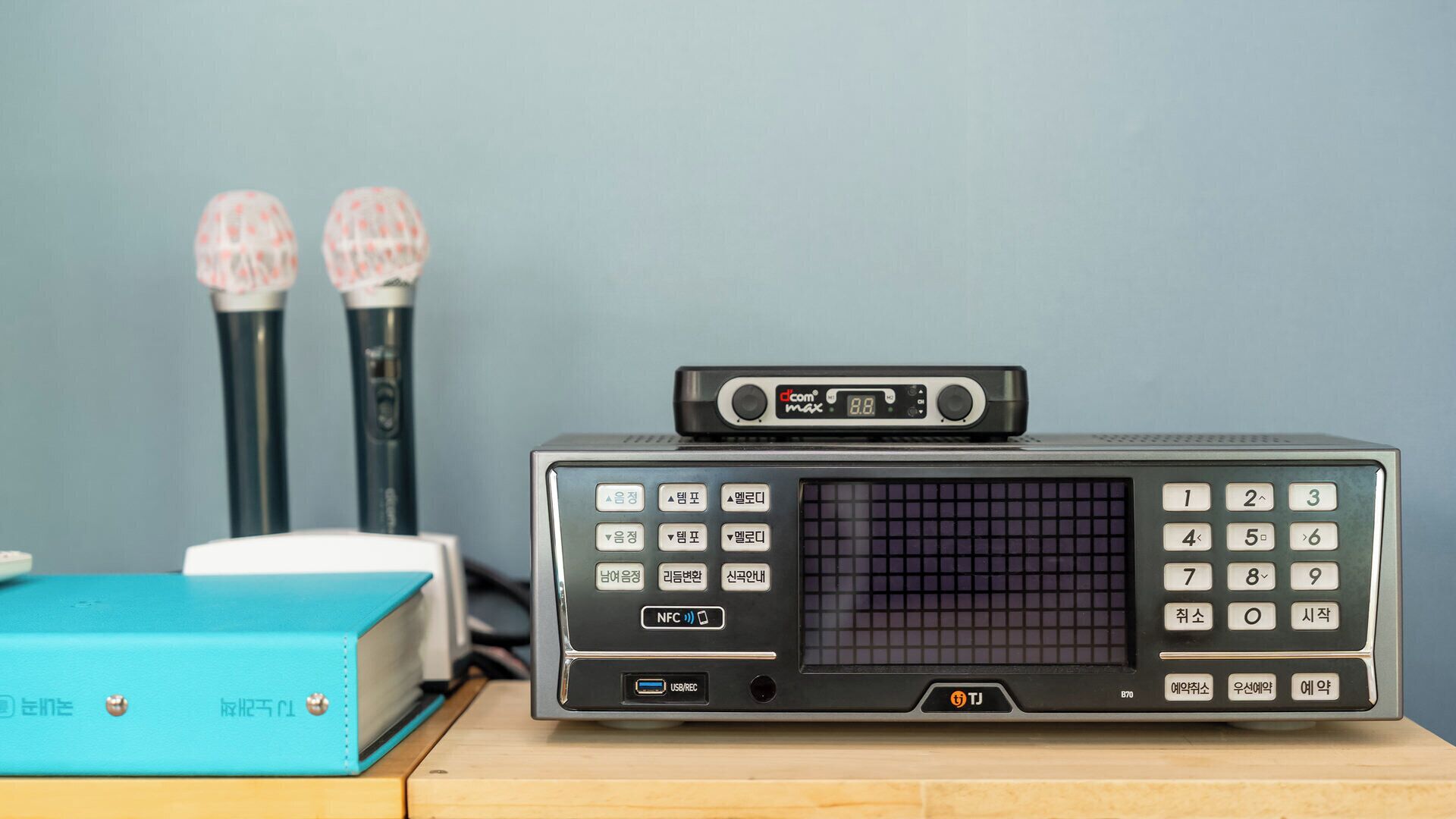 A portable radio with a display, buttons, and a USB port, placed on a wooden surface with a blue box and two cylindrical objects in the background.