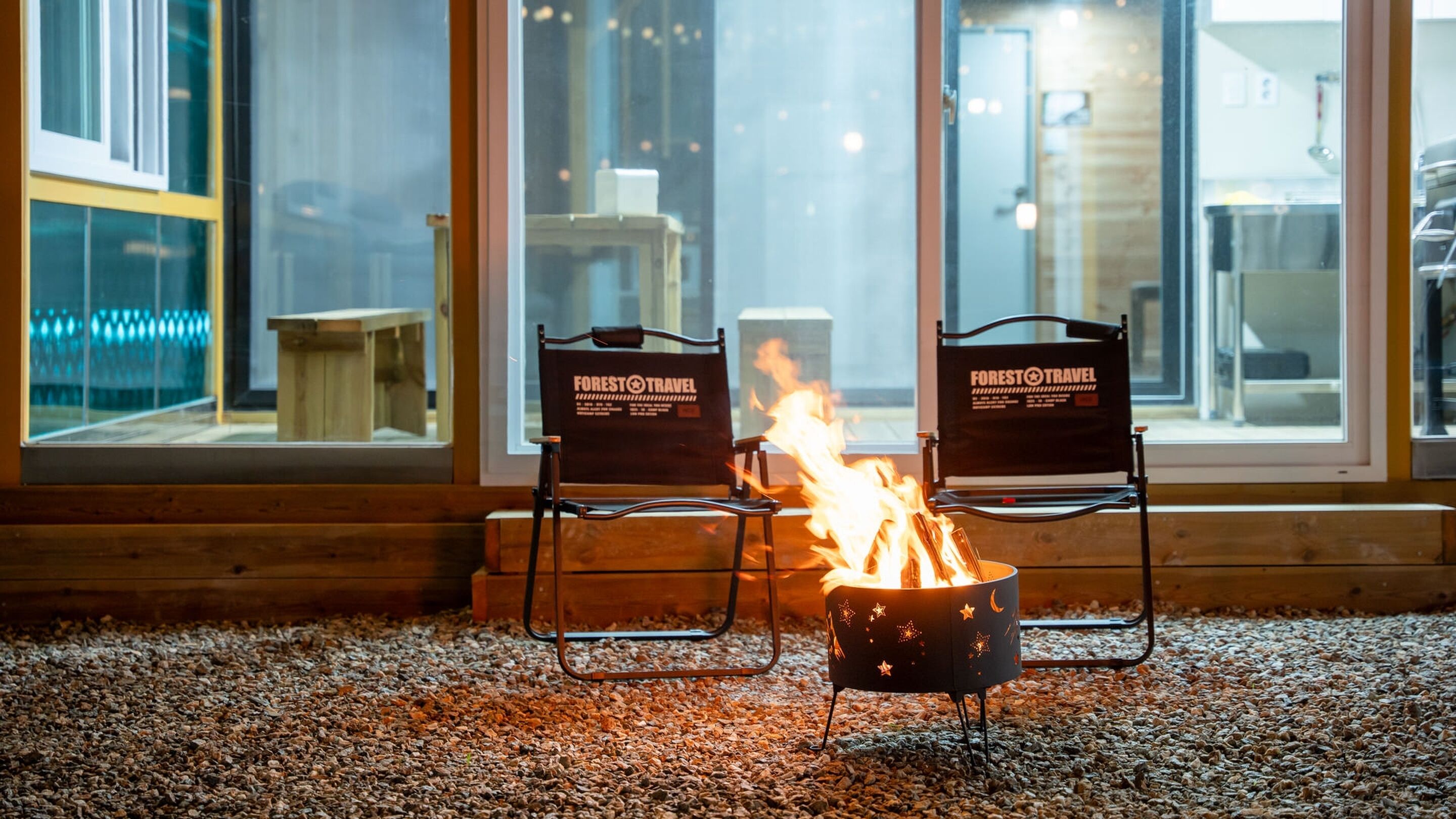 Two chairs with 'FOREST TRAVEL' branding, a small campfire, and a view through large windows.