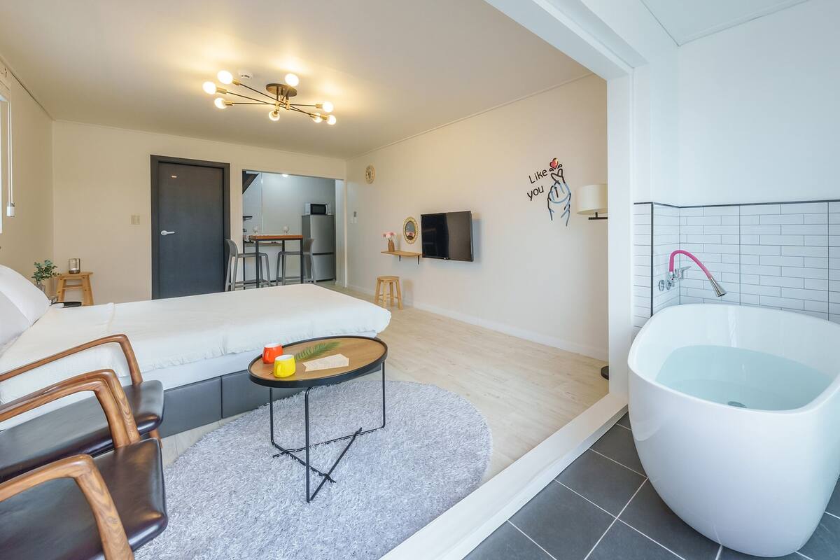 A modern hotel room with a large bathtub, a sofa, a dining area, and a TV.