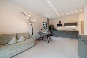 A modern living room with a sofa, dining table, and chairs. A kitchen area is visible in the background with cabinets and a microwave.