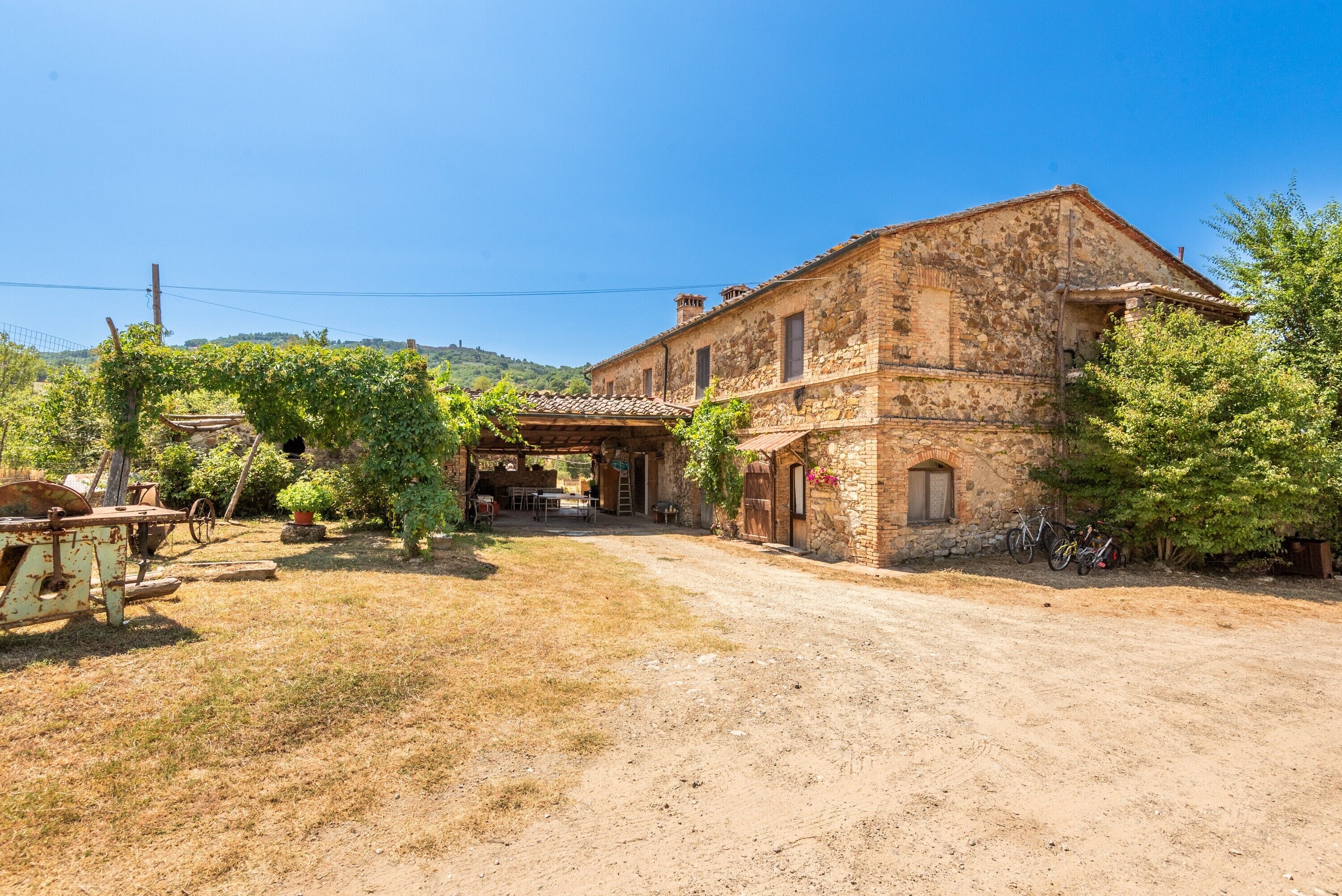 A rustic stone building with a dirt driveway, surrounded by greenery and a clear sky.