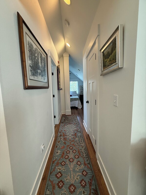A hallway with framed artwork on the walls, a patterned rug, and a bed in the background.