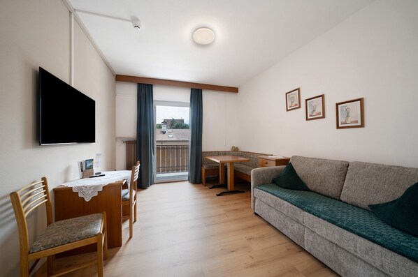 A living room with a flat-screen TV, a sofa, a dining table with chairs, and a balcony with curtains.