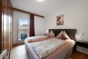 A bedroom with a bed, a nightstand, a wall-mounted lamp, a window with curtains, and a view of buildings outside.