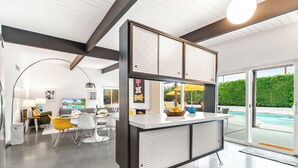 A modern kitchen with a central island, a dining area with chairs, and a view of a swimming pool. - The Butterfly Alexander  – Stylish Mid-Century Home with Pool, Spa, Views & Fire Pit (Palm Springs)