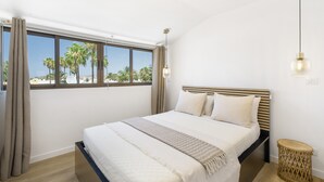 Quarto com uma janela grande, uma cama com cabeceira, um abajur e uma pequena mesa de cabeceira de vime.