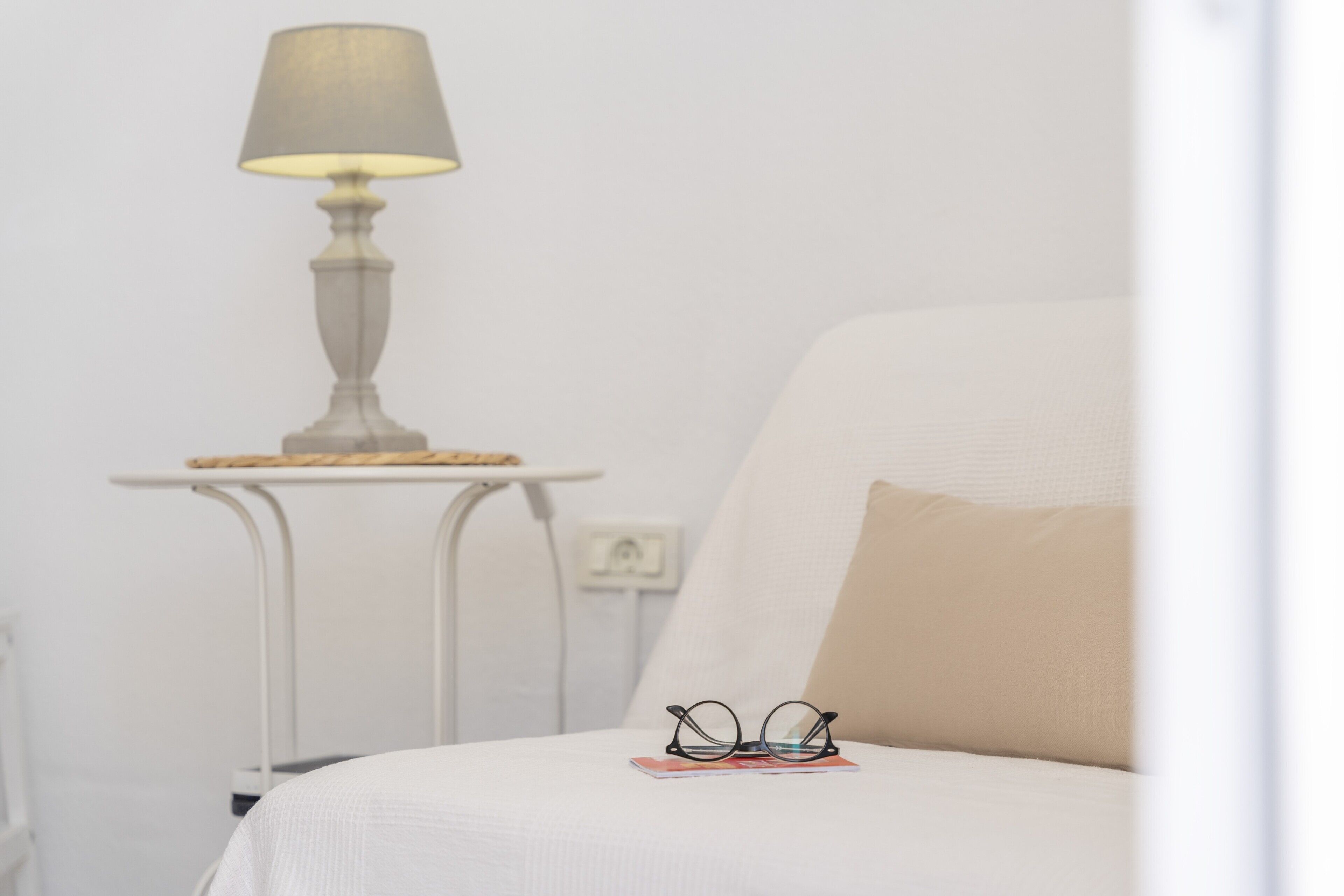 Un lit avec un oreiller, une table de chevet avec une lampe et une paire de lunettes posée sur le lit.