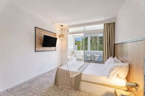 Ein Hotelzimmer mit Bett, Wandfernseher, Balkon mit Tisch und Stühlen sowie Blick auf Grünfläche.