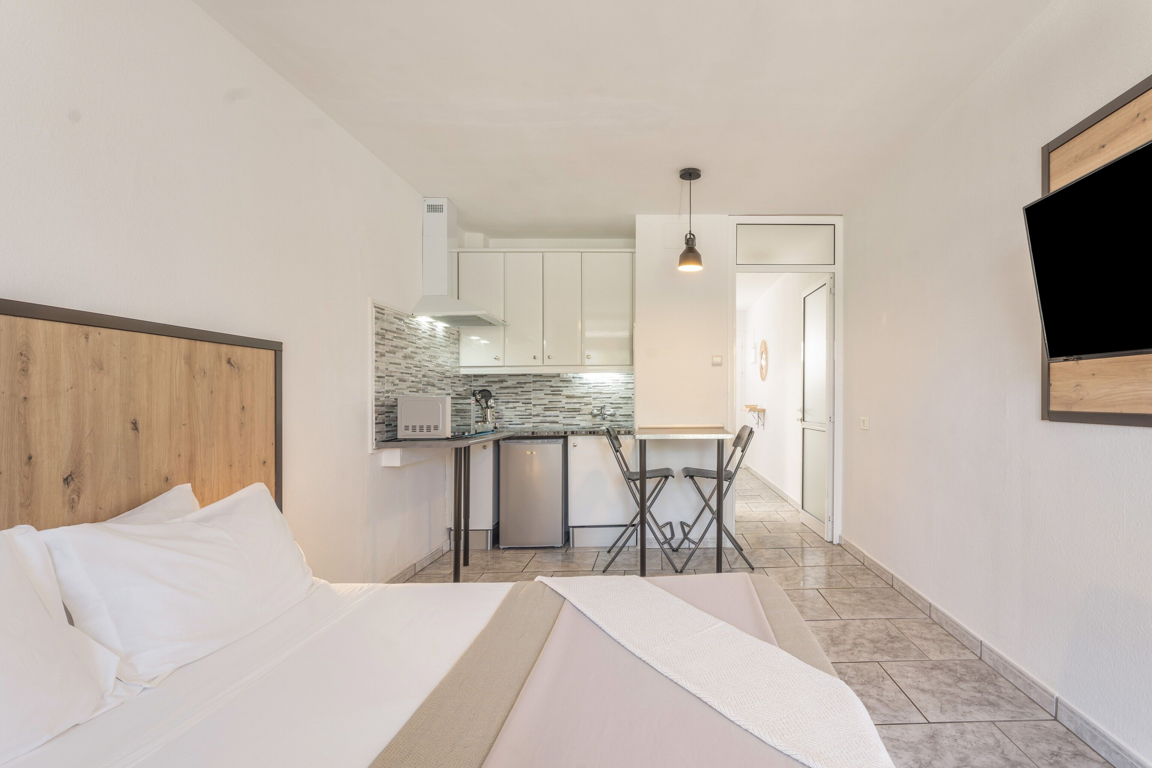 A hotel room with a bed, a small kitchenette, a dining area, and a TV mounted on the wall.