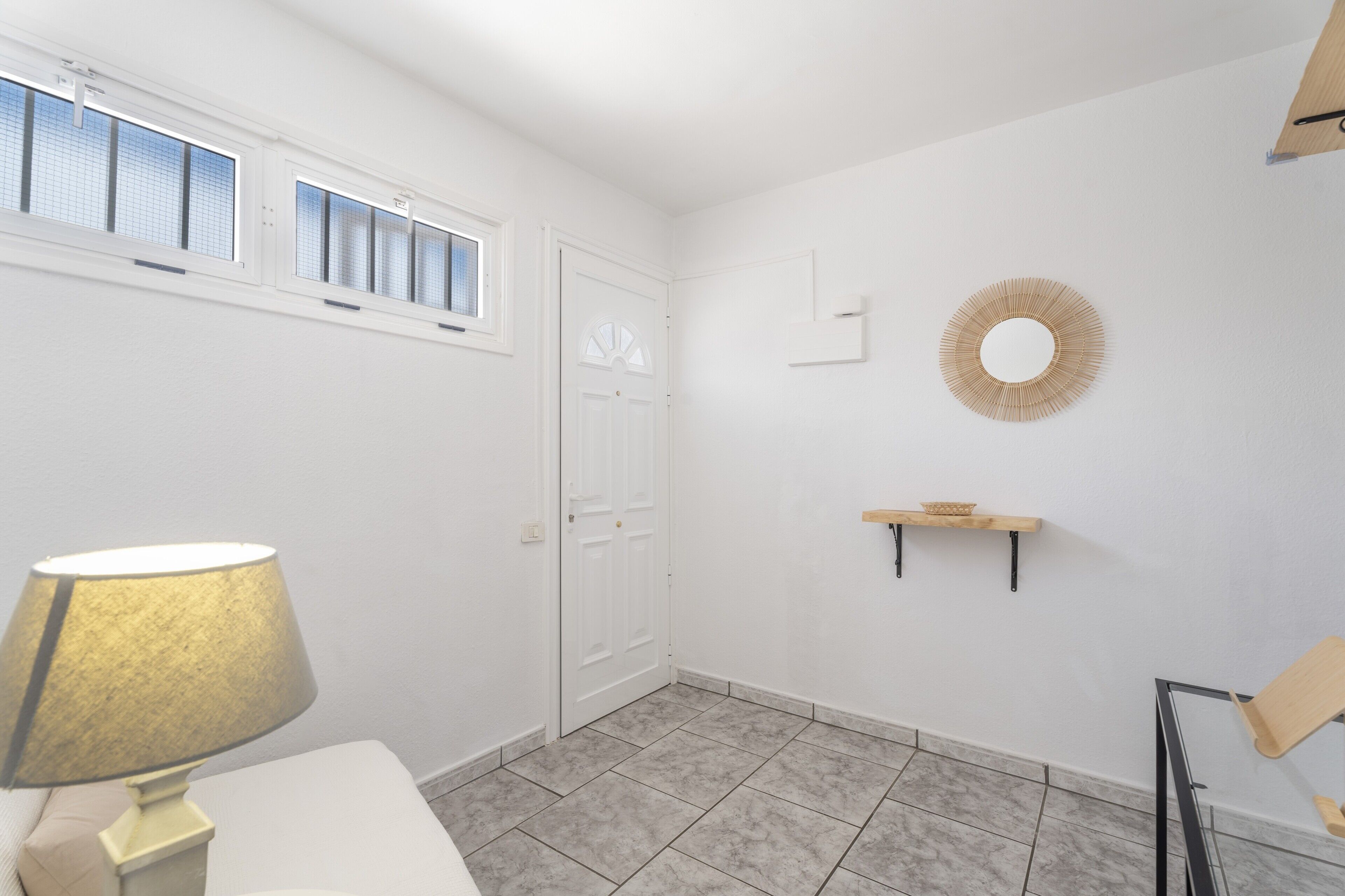 Een kamer met een witte deur, een raam met jaloezieën, een lamp, een wandplank en een ronde decoratie aan de muur.