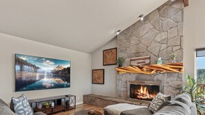 A living room with a stone fireplace, a large TV mounted on the wall, and a cozy seating area with a sofa and cushions. - Downtown Close Lake View with Tesla Charging (Coeur D Alene)
