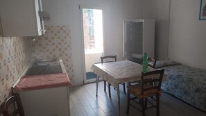 A small kitchen with a sink, a table with chairs, a bed with a floral bedspread, and a window with a view of greenery.