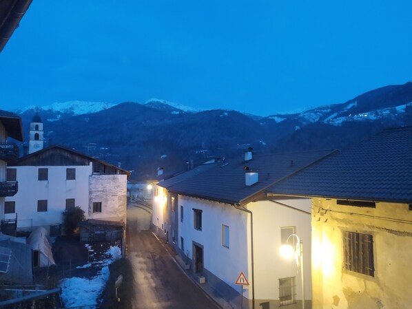 Eine schmale Straße mit Häusern an beiden Seiten, ein schneebedeckter Berg im Hintergrund und ein Straßenschild, das auf eine enge Strecke weist.