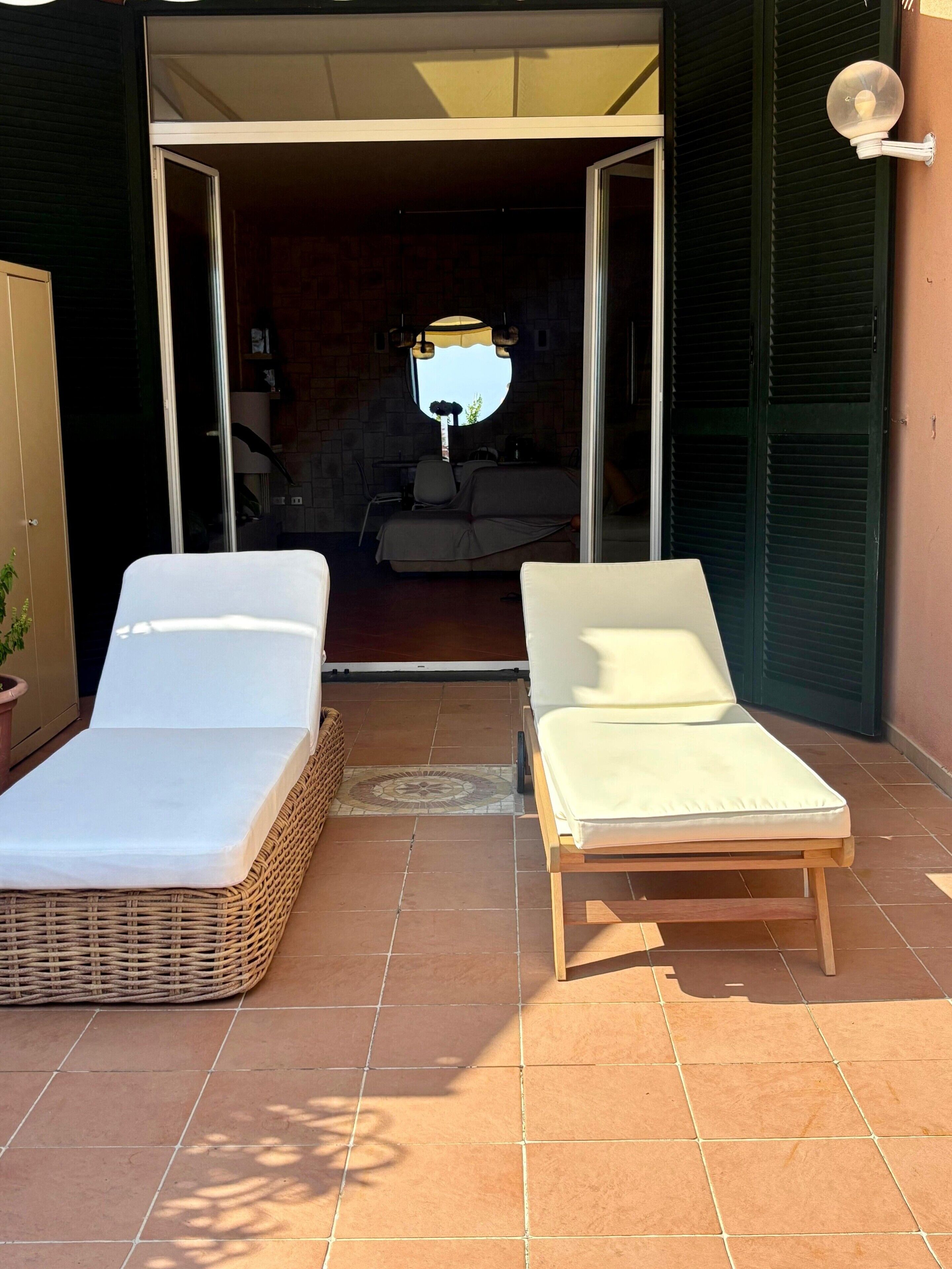 A patio with two sun loungers and a wicker chair, a small table, and a lamp.