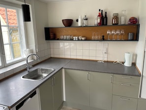A kitchen with a sink, a dish rack, a coffee maker, and a shelf with bottles and glasses.