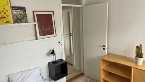 Une chambre à coucher avec un lit, une table de chevet, une lampe, une étagère avec des livres, une porte et une petite plante.