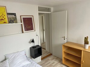 A bedroom with a bed, a bedside table, a lamp, a shelf with books, a door, and a small plant.