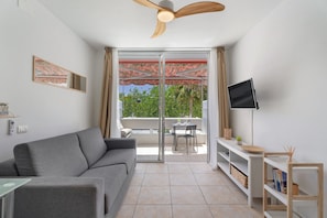A living room with a grey sofa, a TV on a stand, a ceiling fan, and a sliding glass door leading to an outdoor area with a table and chairs. - Vacation Studio 'Greta' with Shared Pool, Balcony, and Wi-Fi (Costa del Silencio)