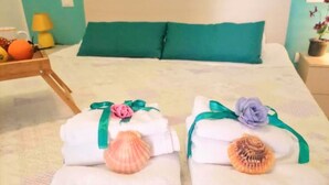 Ein Bett mit zwei Handtüchern, die mit Bändern und Meeresschalen verziert sind, einem türkisfarbenen Kopfkissen sowie einem Nachttisch mit Lampe und Obst.