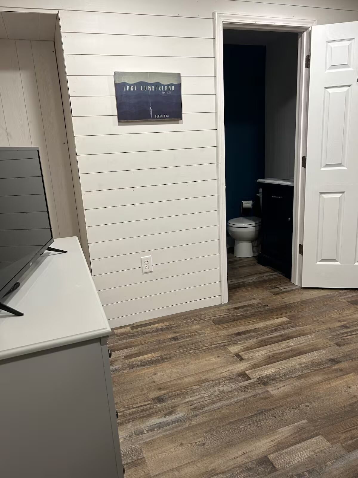 Een kamer met een houten vloer, een wit bureau, een deur naar een badkamer en een bord aan de muur met daarop 'Lake Cumberland'.
