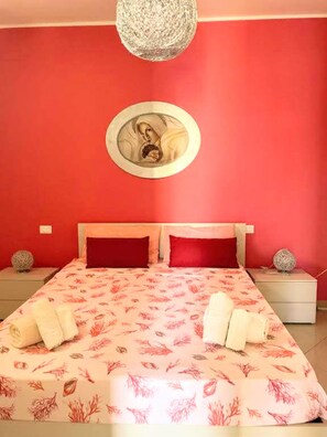 Une chambre avec un mur rouge, un lit recouvert d’une literie à motifs floraux, deux oreillers, un miroir rond et deux tables de chevet ornées d’objets décoratifs.