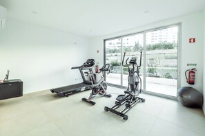 A gym with cardio machines, a weight bench, and a large window with a view of buildings. - Relaxing Pool Stay in Albufeira | Private Terrace (Albufeira)