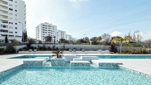 A swimming pool with tiled steps and lounge chairs, surrounded by a fence and buildings.