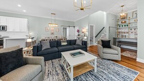 Un vaste espace de vie comprenant un canapé foncé, un fauteuil de couleur claire et une table basse. Un lustre est suspendu au plafond, et des étagères présentent divers objets décoratifs.