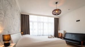 A hotel room with a large bed, a bedside lamp, a black leather sofa, and a window with curtains.