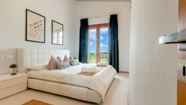 Un dormitorio con una cama, almohadas, una mesita de noche, una lámpara, una ventana con cortinas y dos cuadros en la pared.