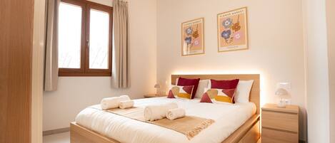 A bedroom with a large bed, two bedside tables, a window with curtains, and two wall-mounted pictures.